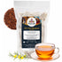 Agulhas Honeybush Tea pouch beside a glass cup of amber tea, loose tea and a yellow flowering sprig.